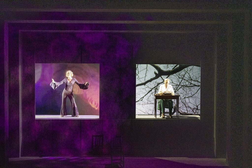 In zwei Vertiefungen auf einer Bühne steht eine Frau in Hosenanzug und ein Mann in Anzug sitzt an einem Tisch.