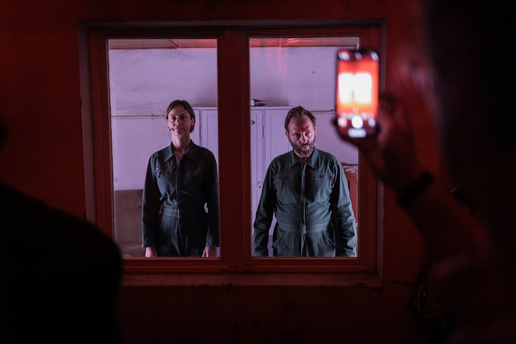 Zwei Menschen in grünen Overalls schauen aus einem Fenster.