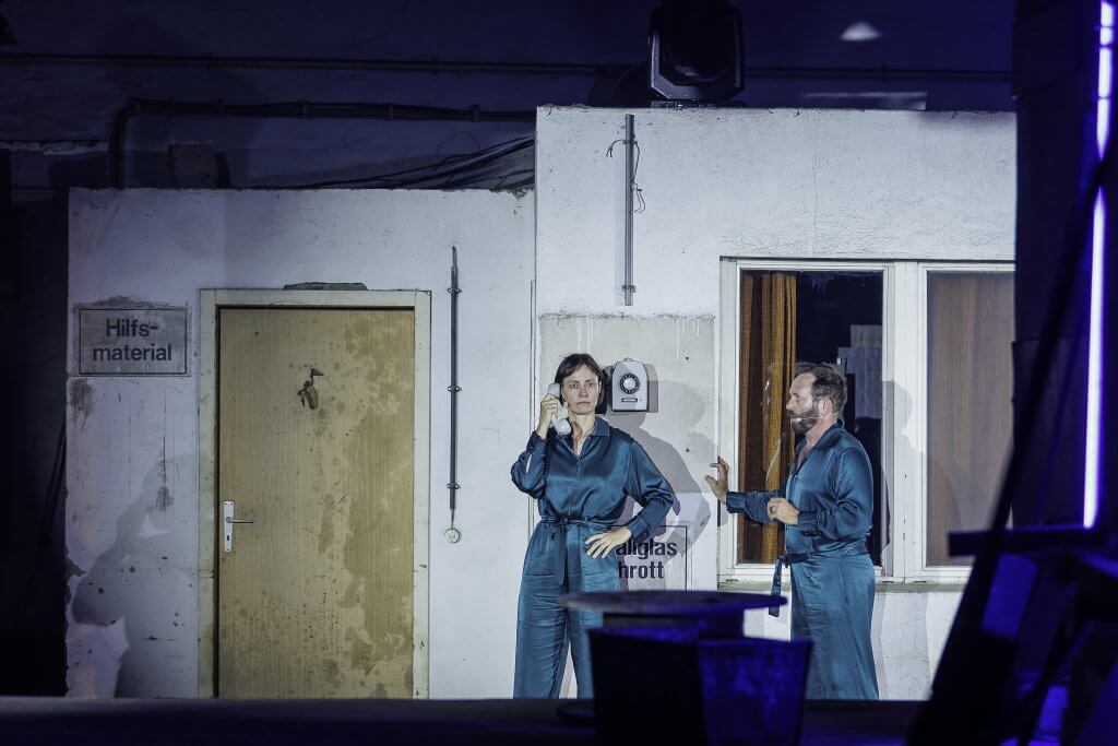 Eine Frau telefoniert mit einem Schnurtelefon an einer weißen Wand. Ein Mann steht daneben und schaut zu.