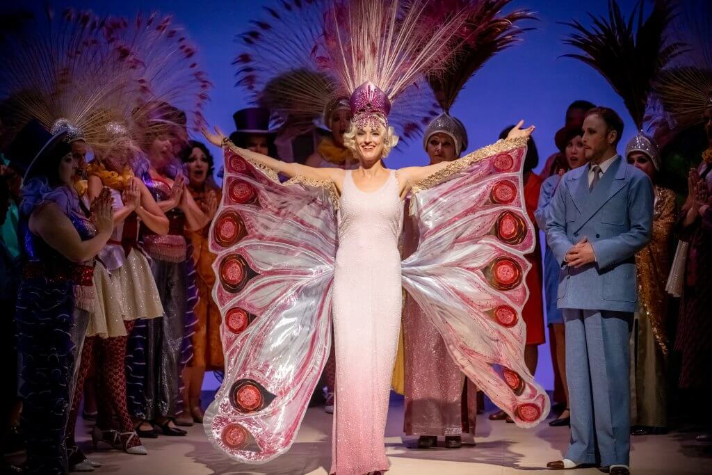 Eine Frau präsentiert ein Show-Outfit mit Schmetterlingsflügeln. Sie ist umringt von zahlreichen Menschen.