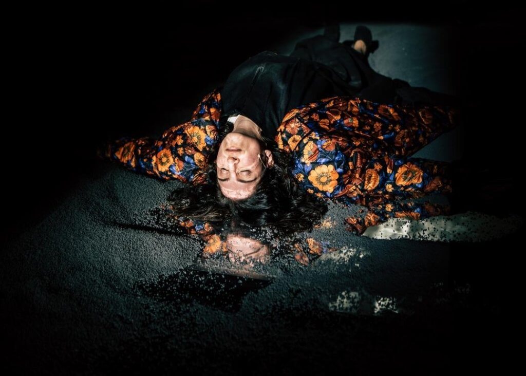 Eine Person mit Blumenjacke liegt auf der Bühne in einer Art Pfütze.