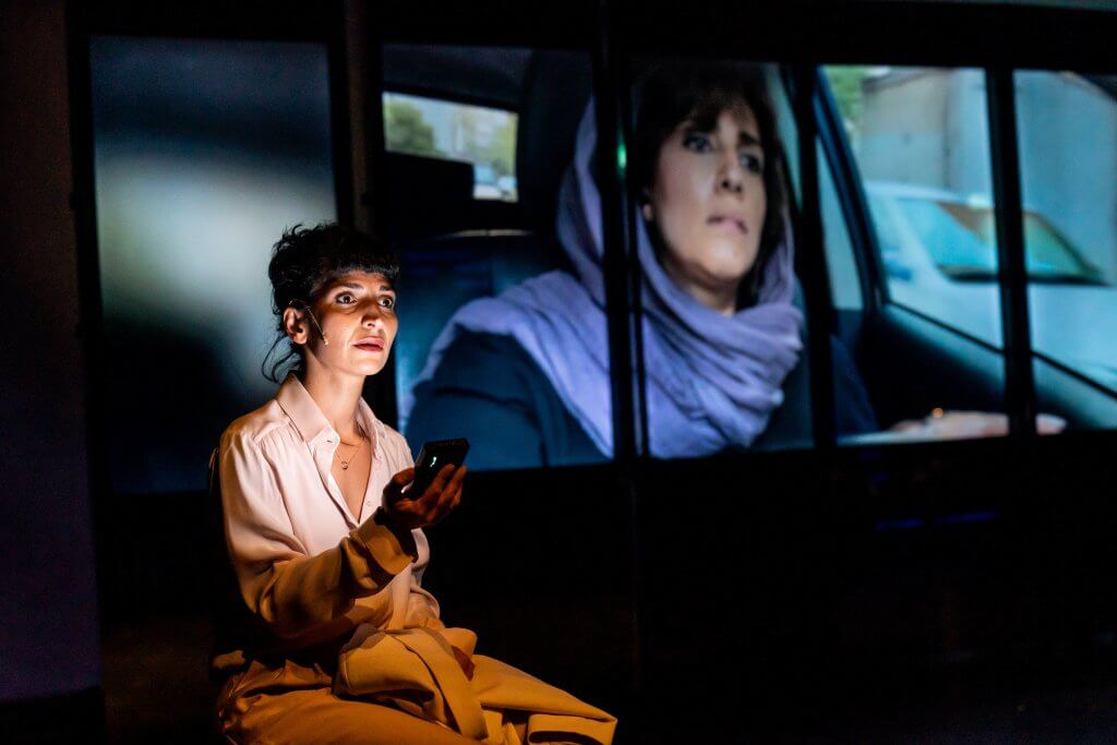 Eine Frau sitzt im Halblicht auf der Bühne. Hinter ihr läuft ein Video einer Frau mit lockerem Kopftuch.