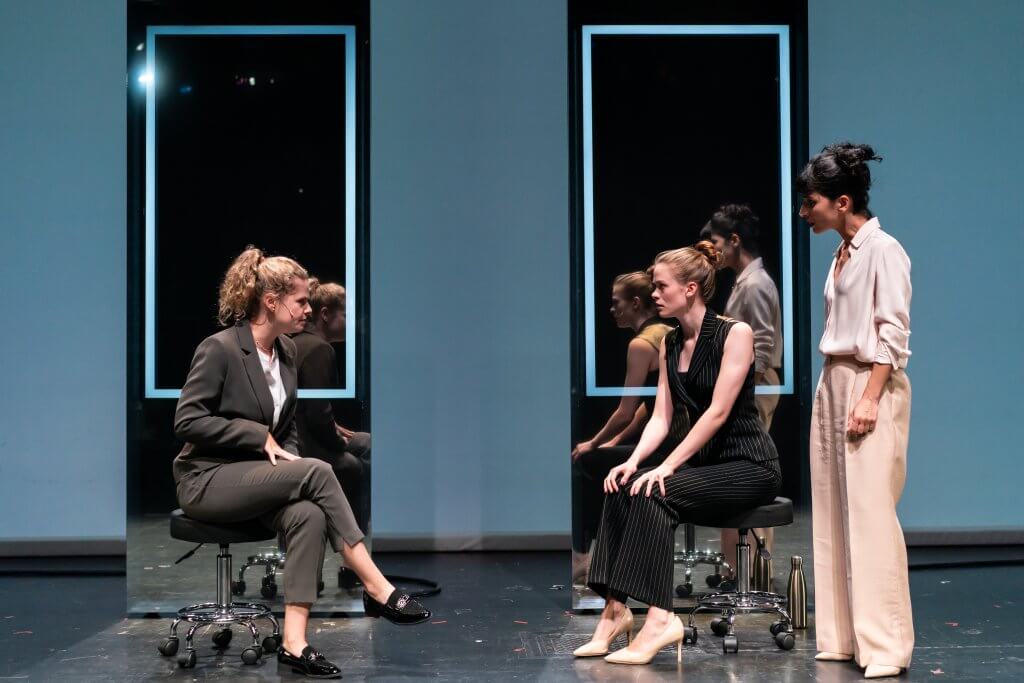 Zwei Frauen sitzen sich gegenüber auf zwei Hocken. rechts steht eine weitere Frau.