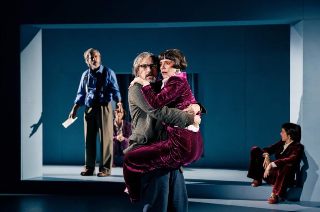 Eine Frau Frau in einem weinroten Kleid klammert sich an einem Mann, der sie hält. im Hintergrund zwei weitere Personen.