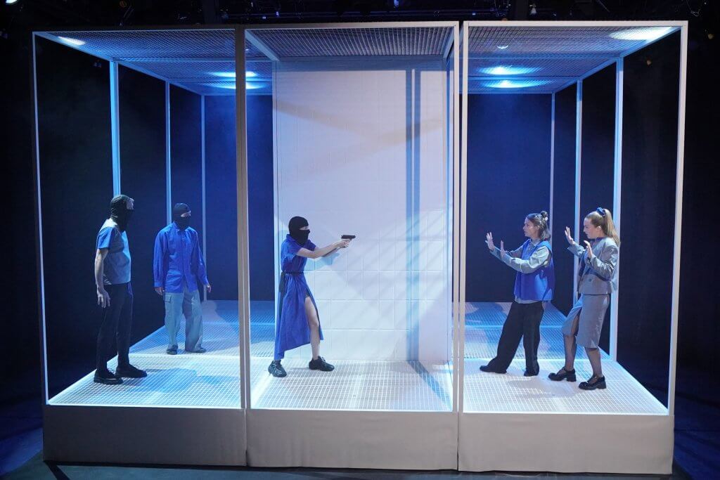 Eine Person mit Sturmmaske bedroht zwei andere Personen mit einer Pistole.