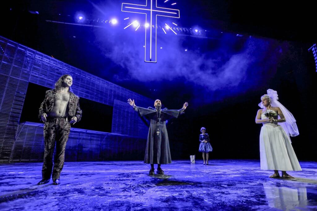 Umringt von einzelnen Menschen steht eine Priesterfigur unter einem blau leuchtenden Kreuz und streckt die Arme zur Seite.