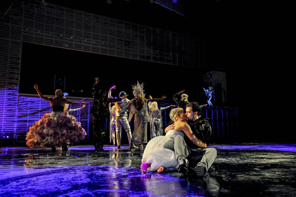 Ein Paar hockt auf dem Bühnenboden und küsst sich. Im Hintergrund feiern mehrere Menschen.