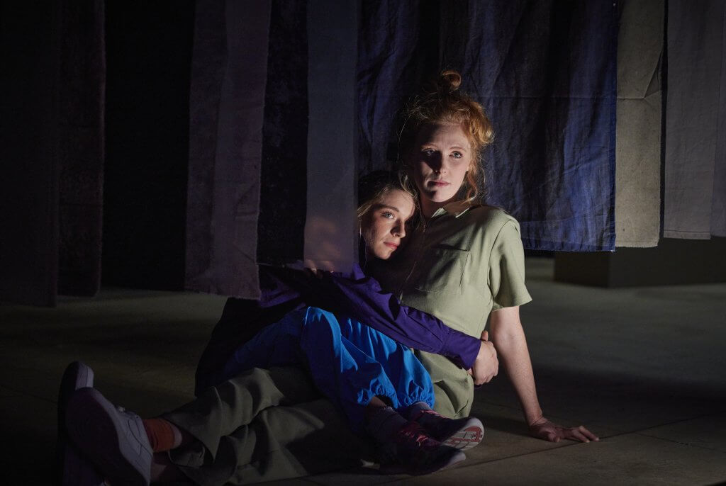 Zwei Personen sitzen zwischen Stoffstreifen, die von der Decke hängen, auf dem Boden und klammer sich aneinander.