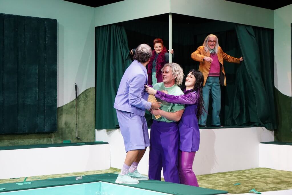 Drei Personen halten sich aneinander, im Hintergrund treten zwei Personen hiner Vorhängen hervor.