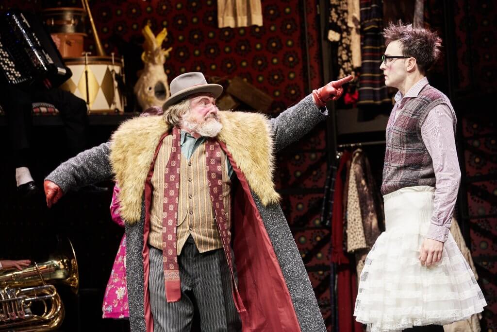 Ein Mann mit breitkrempigen Hut zielt demonstrativ auf den Kopf eines Mannes in Kellner-Outfit.