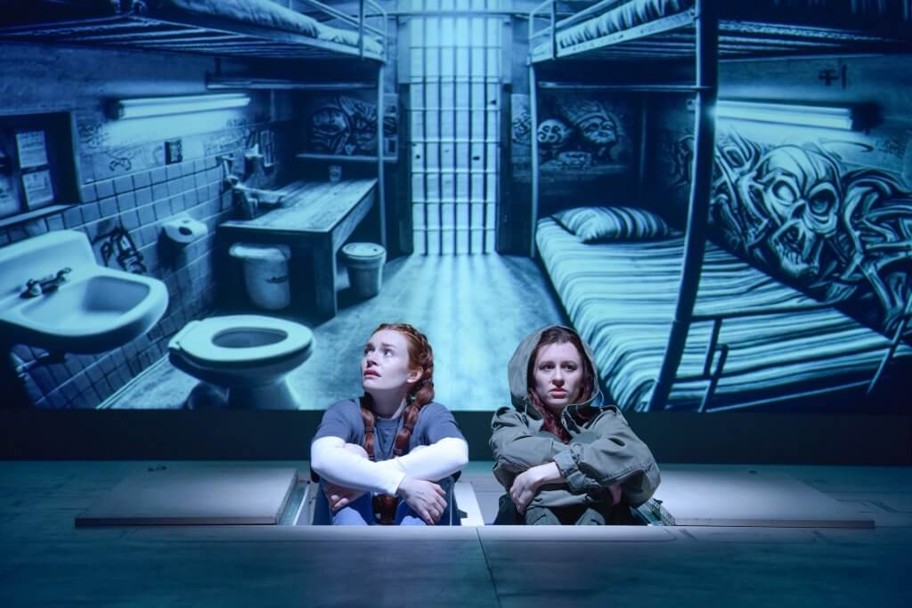 Zwei Frauen hocken zusammengekauert nebeneinander vor der Projektion einer Gefängniszelle.