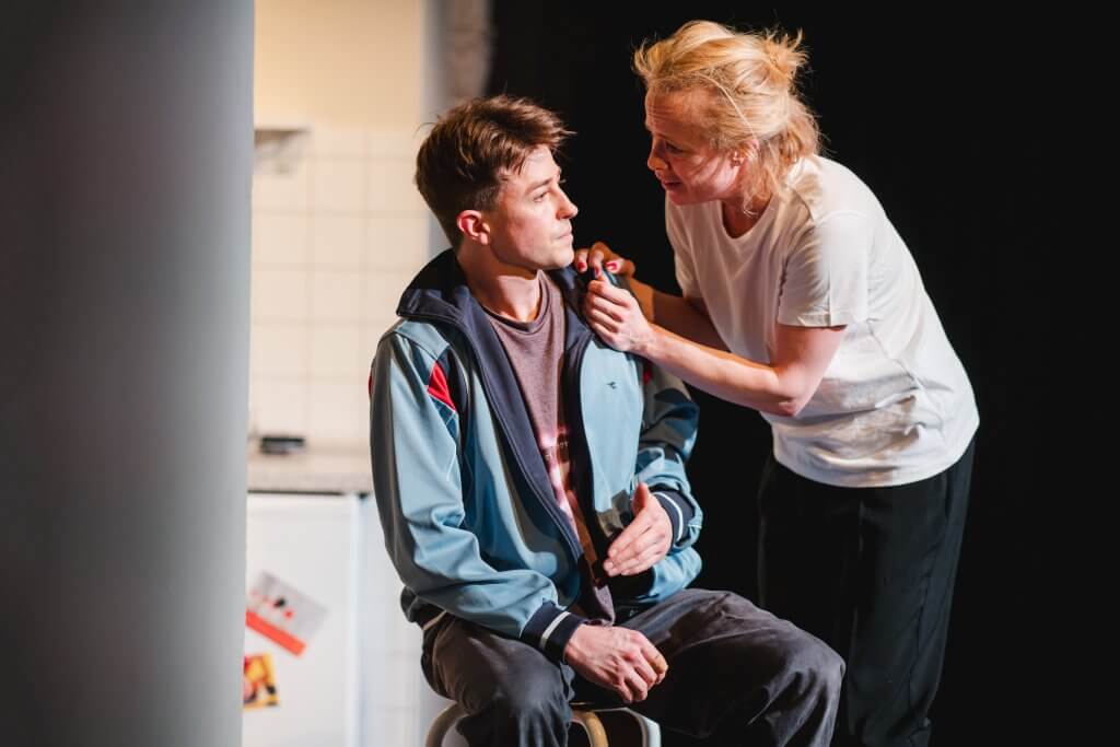 Ein Junge sitzt auf einem Stuhl, eine Frau mit blonden Haaren fast ihn emotional an der Schulter an.