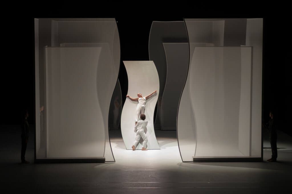 Durchscheinende Wände mit geschwungenen Kanten, in der Mitte hebt an einer solchen Wand ein Mann eine Frau in Weiß hoch.