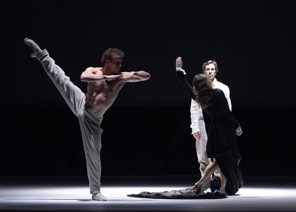 Ein Mann in grauer Hose streckt sein Bein nach hinten oben. Eine Gestalt im dunklen Kleid geht an einer weiteren Person in Weiß vorbei.