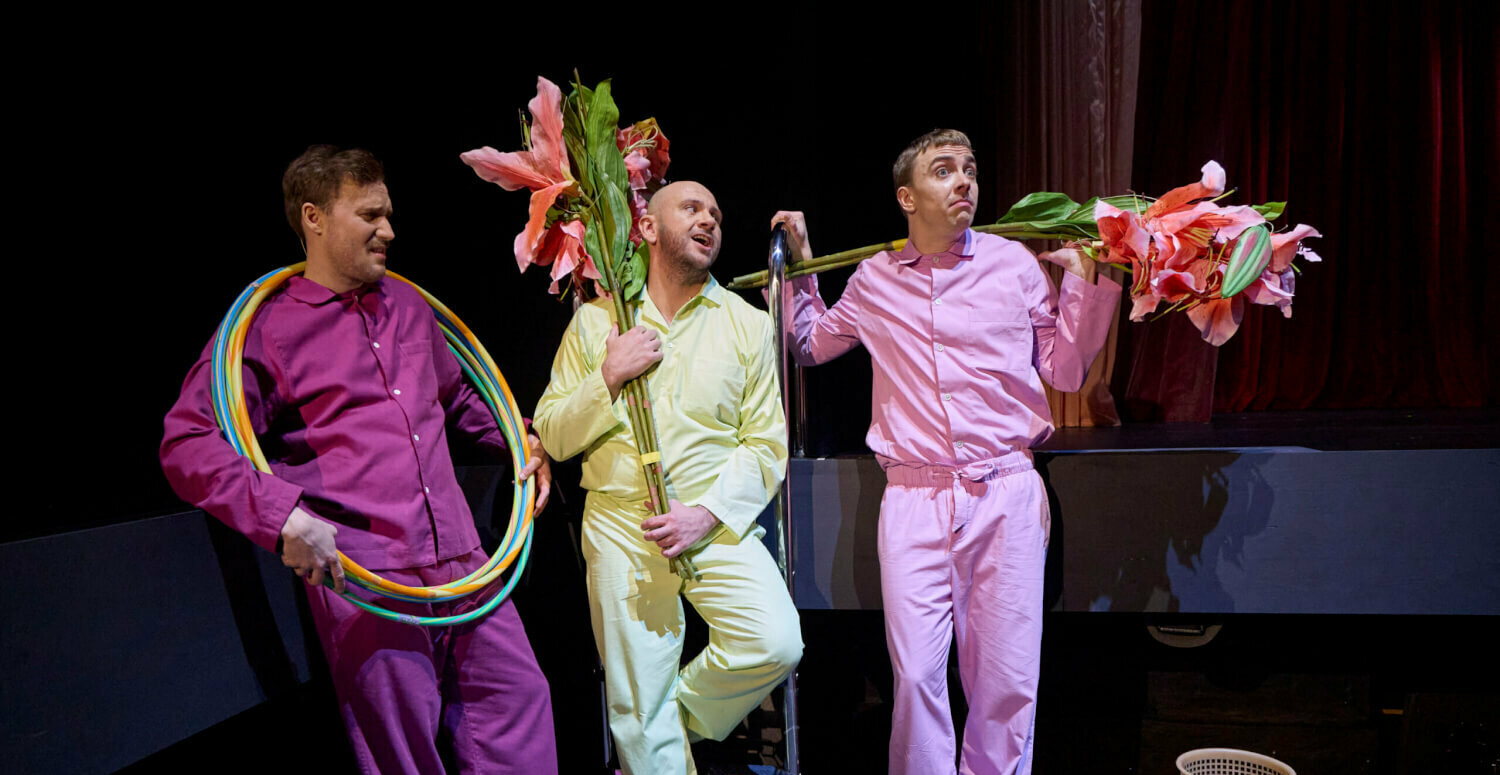 drei männlich gelesene Personen in Lila, Hellgelb und Rosa mit großen Blumen und Hula-Hoop-Reifen