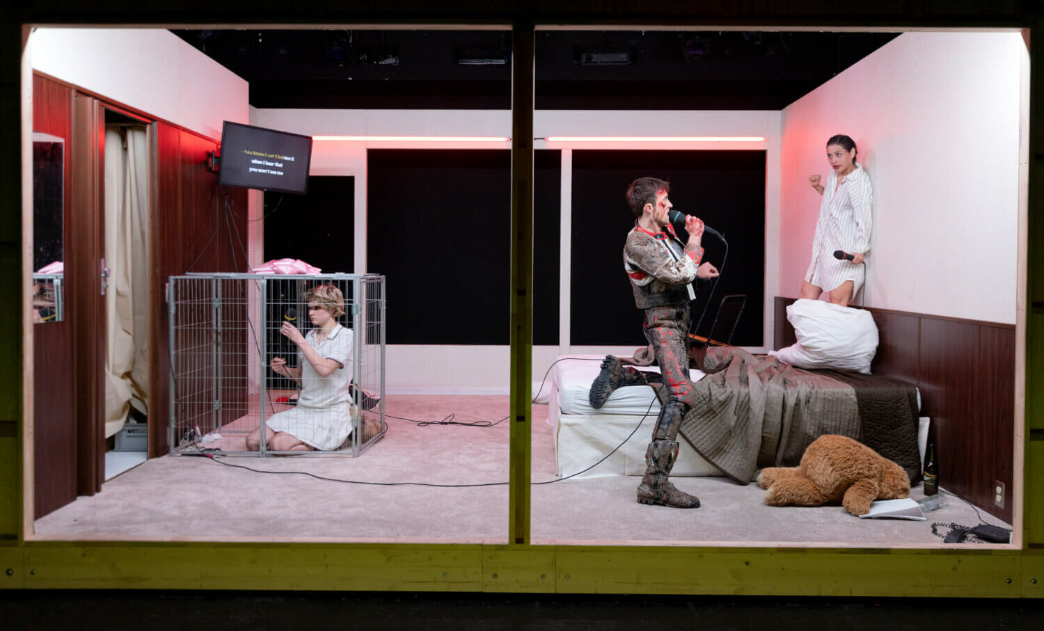 eine Bühne, links kniet eine Person nach links gewandt, rechts eine Person mit Mikro und auf einem Bett eine weitere Person