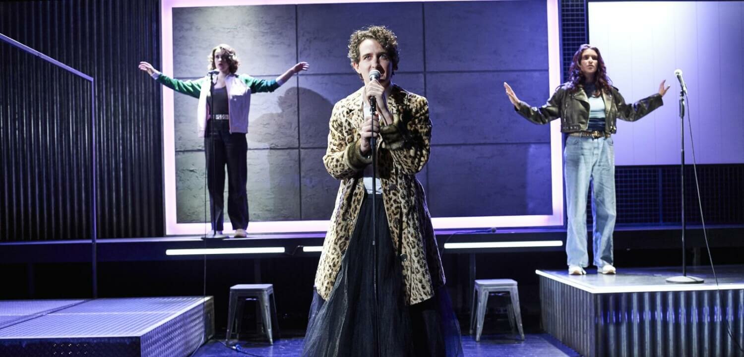 im Vordergrund Person in Leopardenmantel vor Mikro, rechts und links im Hintergrund noch zwei Personen