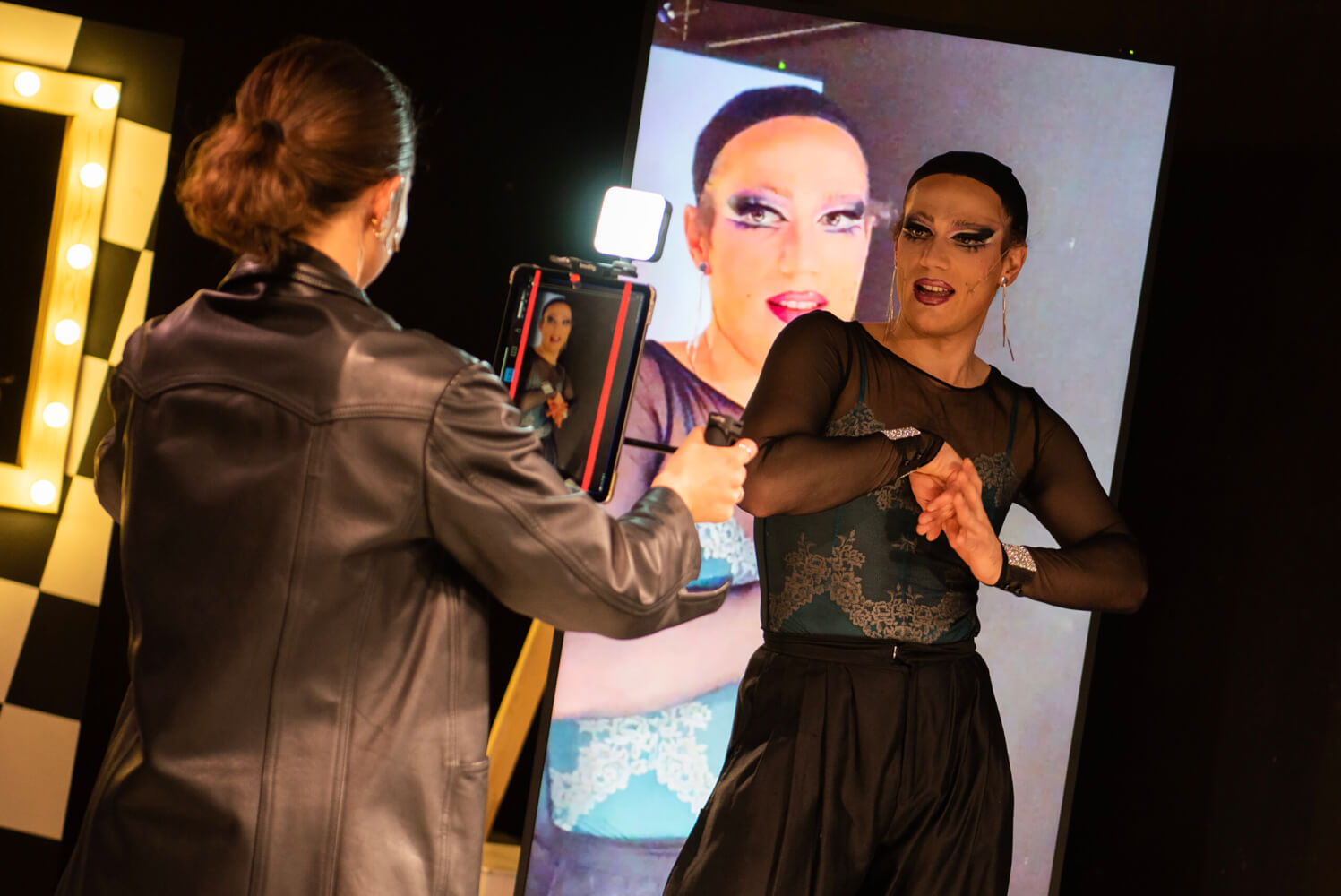 rechts eine darstellende Person, die von vorne links mit einer Kamera aufgenommen wird. im Hintergrund wir das Aufgenommene projiziert