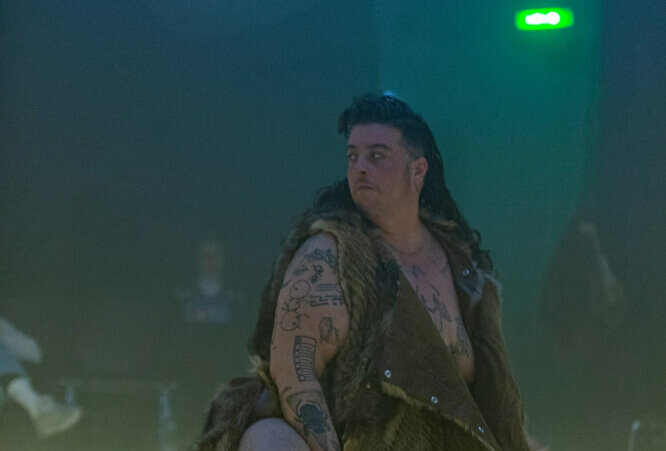 eine Person in einer Art Fell-Kostüm-Mantel steht nach rechts gewandt und schaut über die rechte Schulter zum linken Bildrand