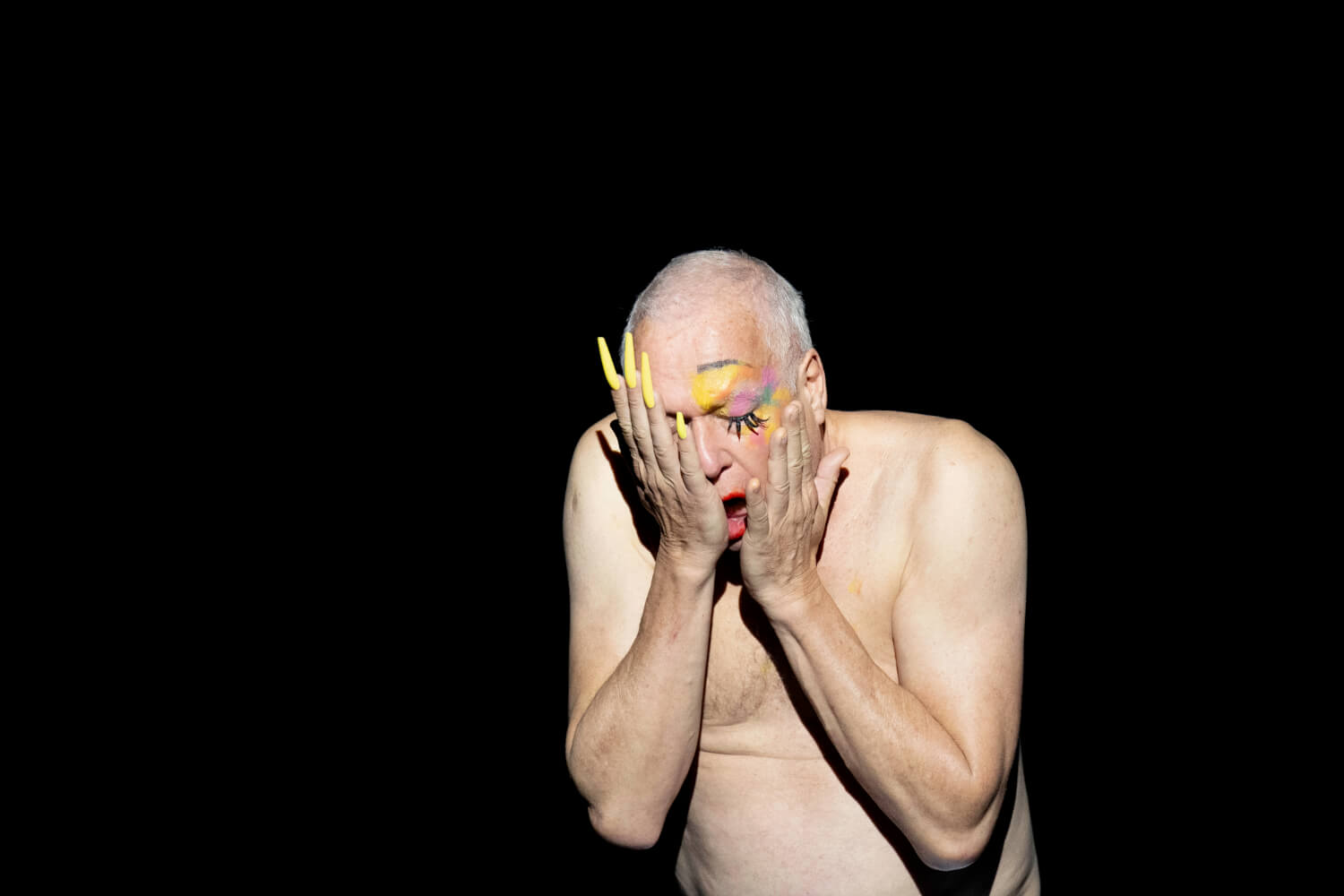 Bariton Johannes Martin Kränzle steht mit nacktem Oberkörper auf der Bühne. Seine Hände sind auf sein Gesicht gelegt. Die linke Hand zieren lange gelbe Fingernägel. Seine rechte Gesichtshälfte wird durch buntes Make-up hervorgehoben.