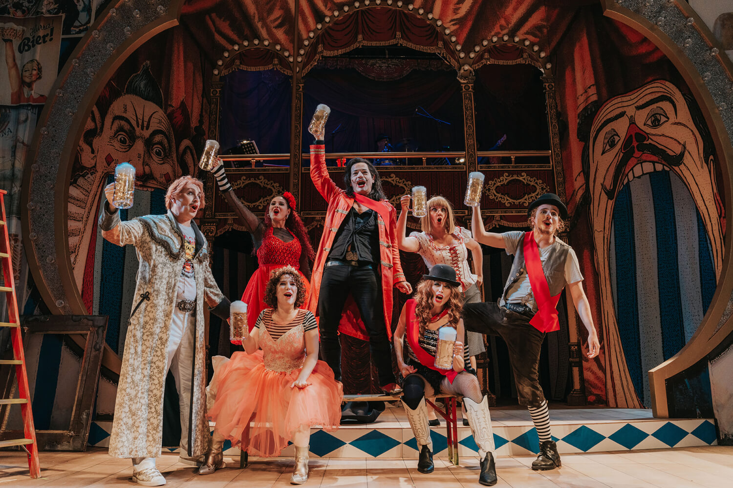 Das Ensemble stößt auf der Bühne gemeinsam an. Alle sind in bunte Kostüme gekleidet.