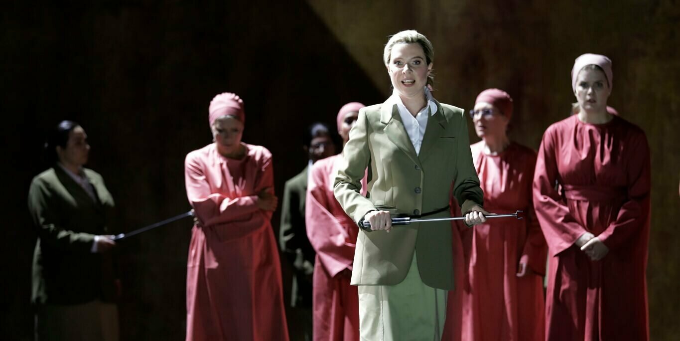 Eine Sängerin im Vordergrund mit Gerte in der Hand, dahinter mehrere Personen in roten Gewändern