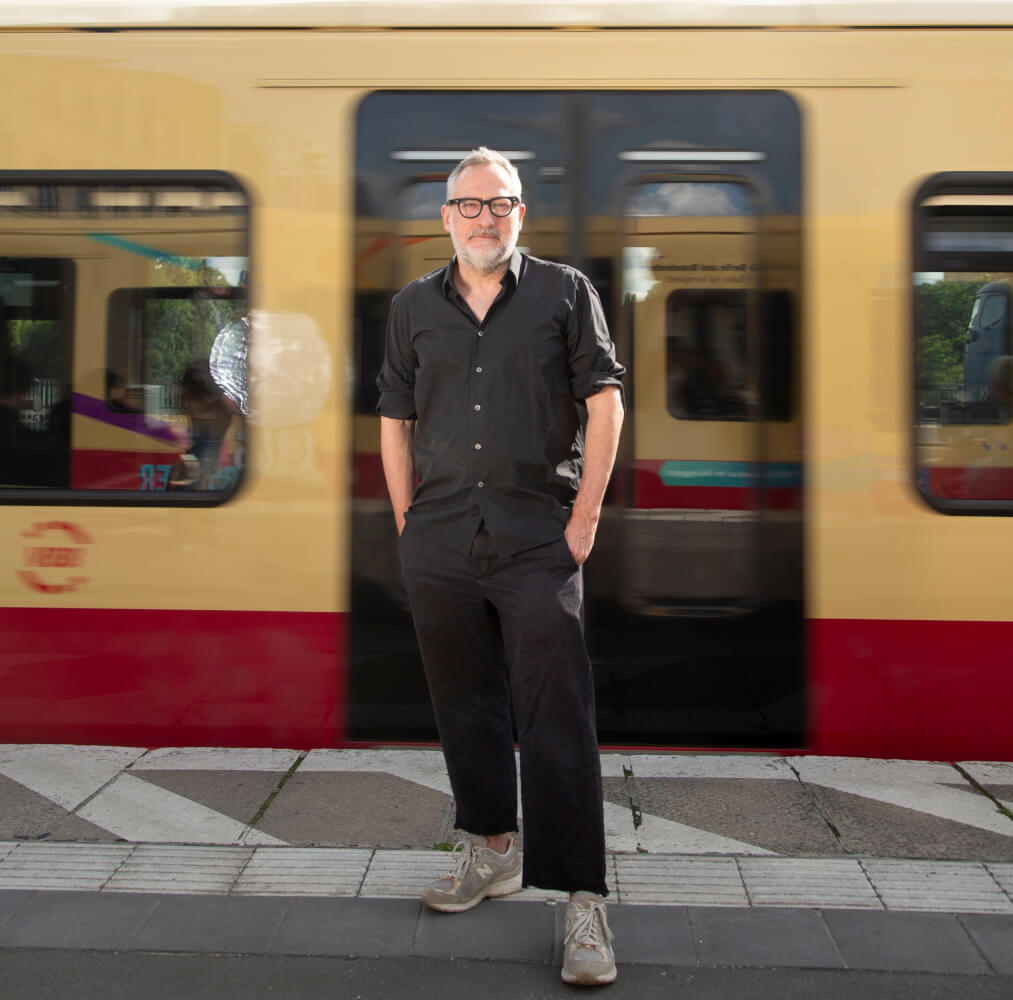 Zu sehen ist der Autor Roland Schimmelpfennig auf einem Bahnsteig. Dicht hinter ihm fährt eine Berliner S-Bahn an ihm vorbei.