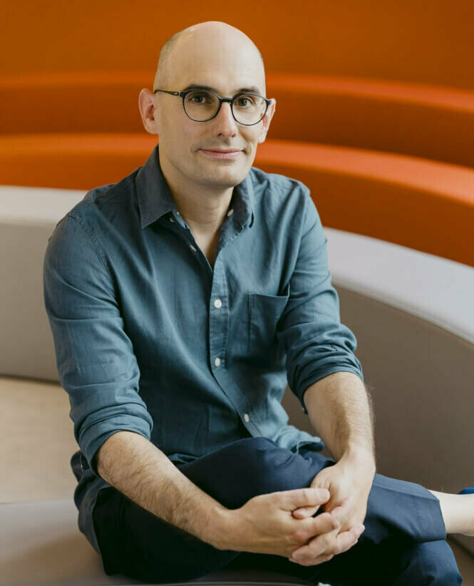 Zu sehen ist Alexander Riemenschneider, Intendant des Theater an der Parkaue, sitzend, die Hände umschlingen das Knie