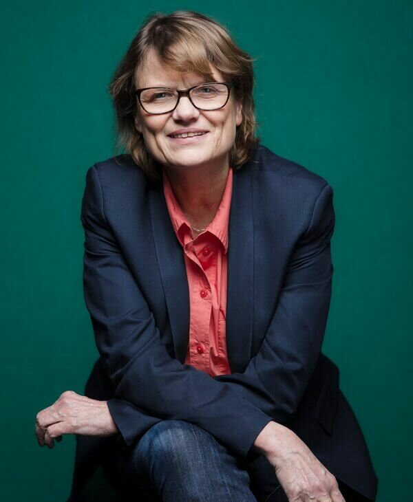 Zu sehen ist Nicola May, Intendantin des Theater Baden-Baden, vor einem grünen Hintergrund, mit überschlagenen Beinen und Armen
