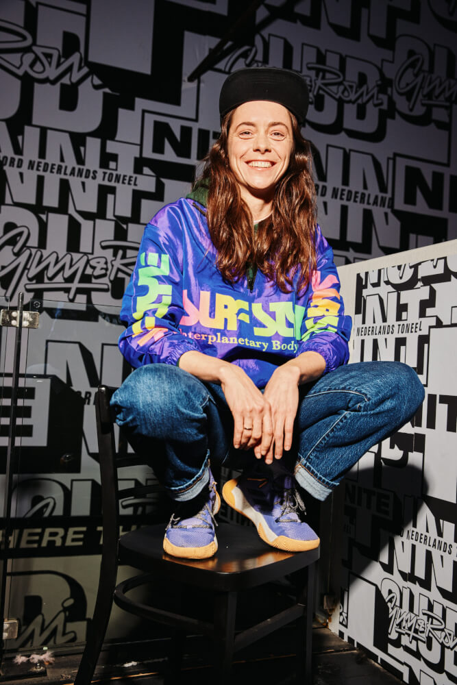 Zu sehen ist die Dramaturgin Friederike Schubert auf einem Stuhl hockend und in die Kamera lächelnd