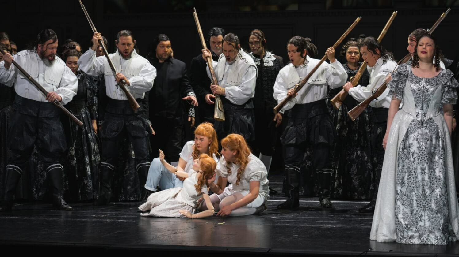 Der Opernchor umzingelt zwei Mädchen und eine Puppe, die in seiner Mitte auf dem Boden sitzen und richtet Waffen auf sie.