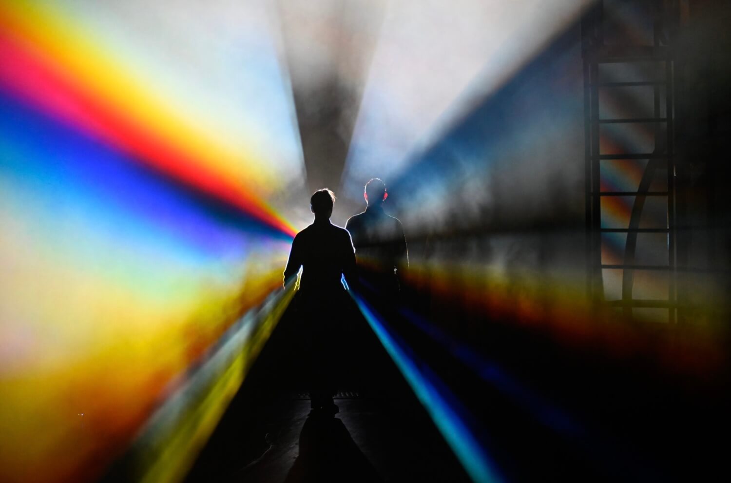 zwei Silhouetten vor schwarzem Hintergrund mit regenbogenfarbener Strahlung