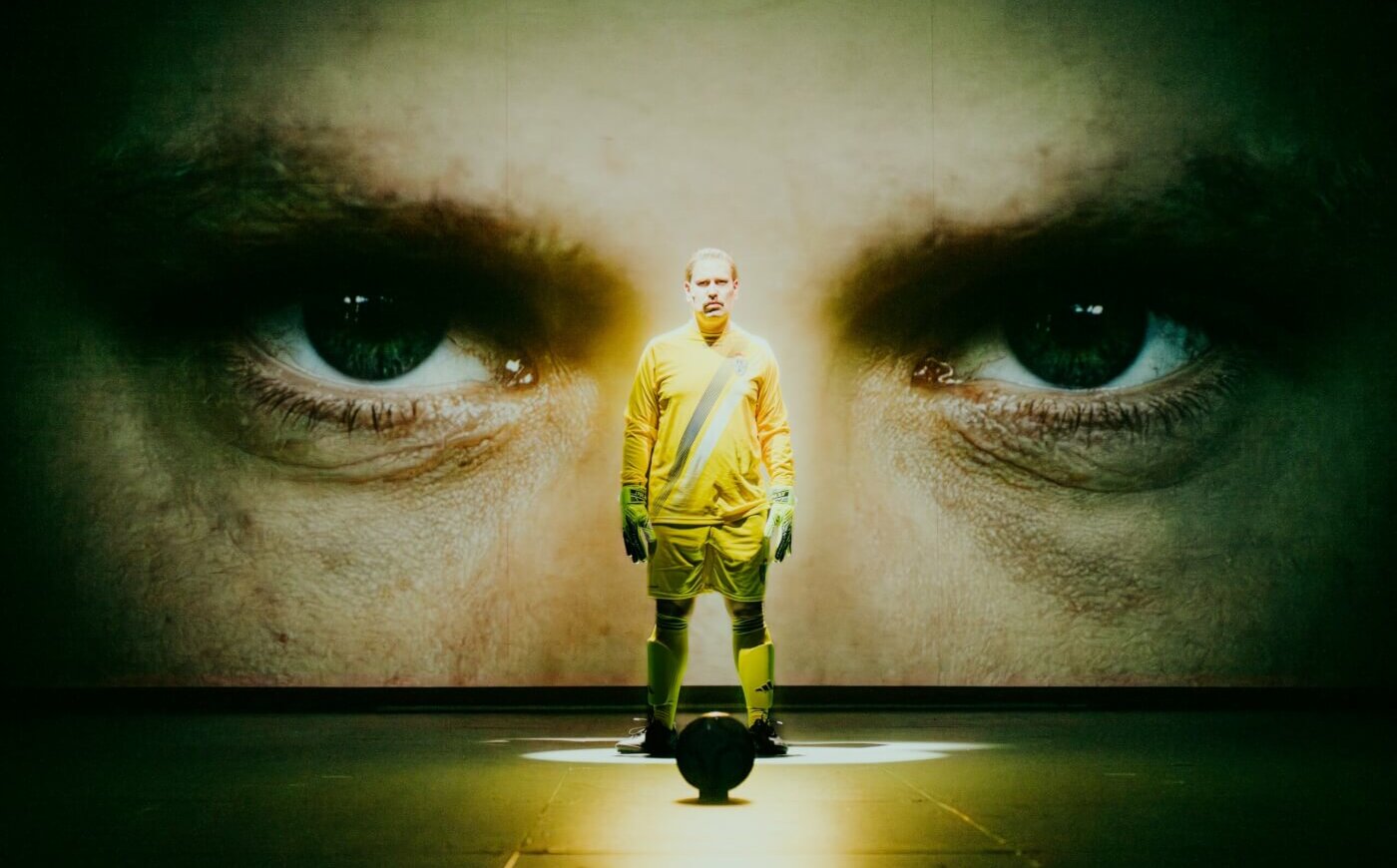Schauspieler im Fußballtrikot vor Rückwand mit seiner projizierten Augenpartie