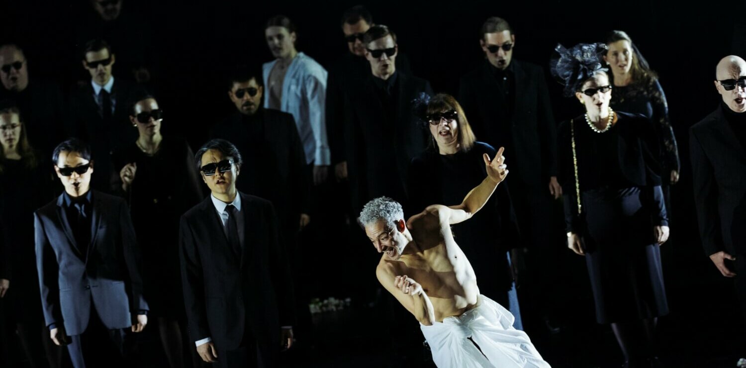 Der Opernchor steht in schwarzer Abendgarderobe und Sonnenbrillen im Hintergrund. Vorne steht ein Mann, um die Hüfte ein weißes Tuch gehüllt.