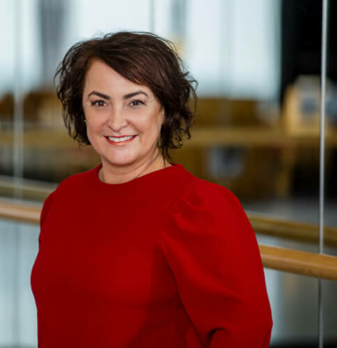 Man sieht Adriana Almeida Pees, Küntlerische Leiterin Tanz am Theater Freiburg. Sie trägt ein rotes Oberteil vor einer Ballettstange und Spiegelwand.