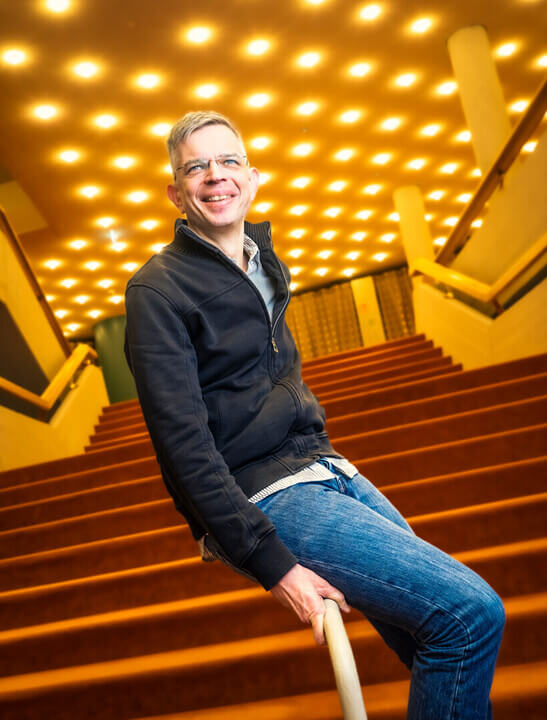 Zu sehen ist Andreas Kornacki, kaufmännischer Geschäftsführer des Düsseldorfer Schauspielhauses, auf einem Treppenlauf sitzend. Hinter ihm sind rote Treppenstufen zu sehen.