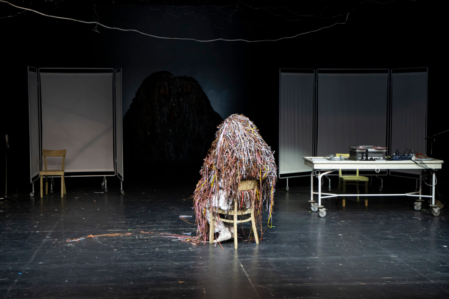 Zu sehen ist ein Szenenfoto aus „Ich, Antonin Artaud – Le Mômo“ (UA) von Roberto Ciulli nach Antonin Artaud. In der Mitte sitzt jemand auf einem Stuhl, überdeckt mit dünnen Fäden.