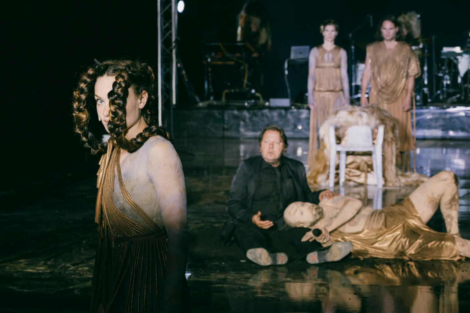 Zu sehen ist ein Szenenfoto aus Philipp Preuss’ Open-Air-Inszenierung von Euripides’ „Backchen“. Im Vordergrund links eine Frau mit zwei großen, geschwungenen Zöpfen. Im Hintergrund zwei Frauen hinter einem Stuhl und davor ein Mann, der auf dem Schoß eines anderen sitzenden Mannes liegt.