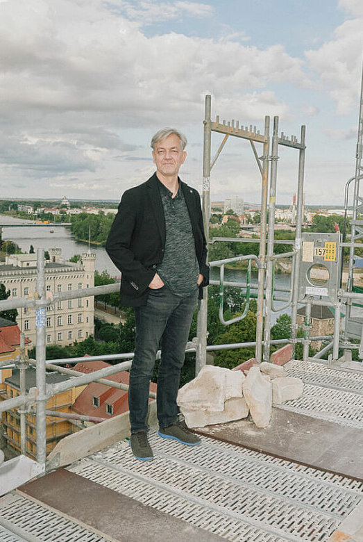 Man sieht Jörg Mannes, Ballettdirektor am Theater Magdeburg, auf einem Baugerüst stehend. Im Hintergrund sind ein Fluss und Häuser von oben zu sehen.