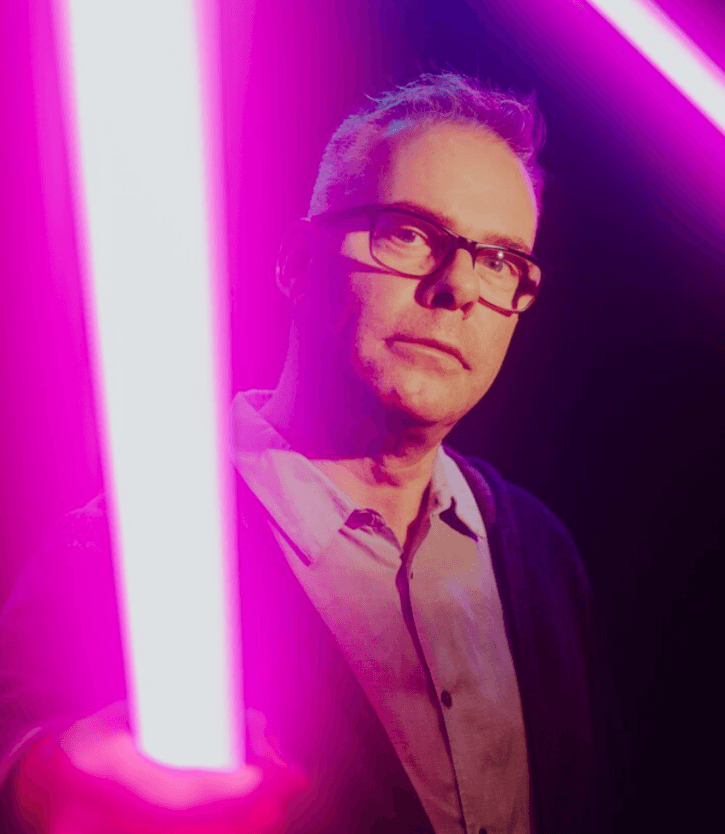 Zu sehen ist Marcus Lobbes, Direktor der Akademie für Theater und Digitalität am Theater Dortmund. Links vor ihm und rechts über ihm leuchten zwei punkfarbene Neonröhren.