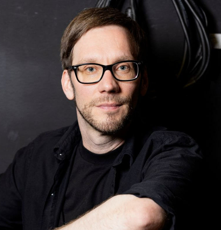 Zu sehen ist Michael Kaiser, Leiter des Jungen Theaters am Theater Freiburg. Er trägt ein schwarzes Oberteil, eine Brille mit schwarzer Fassung vor einem schwarzen Hintergrund.