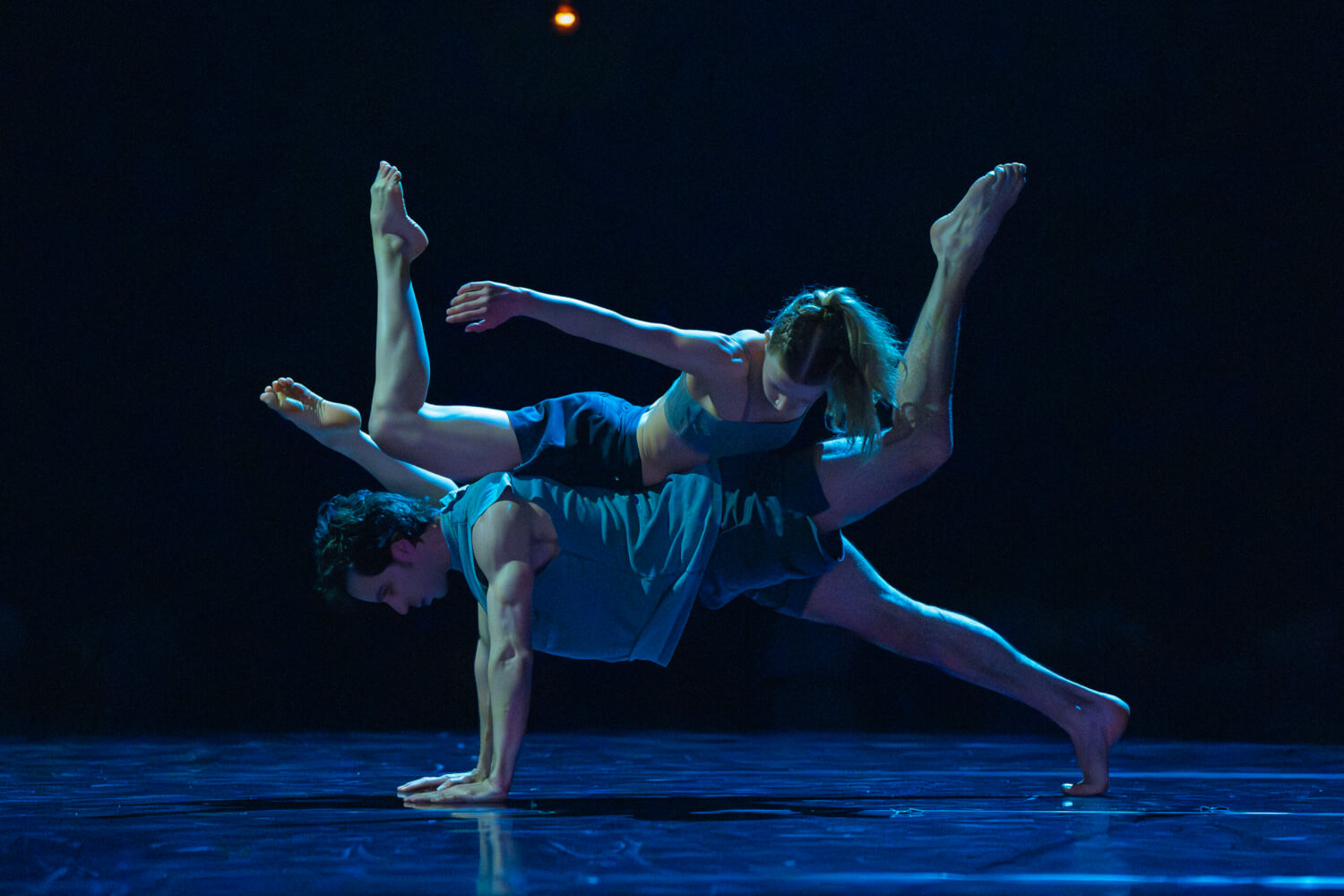Ein Tänzer befindet sich im verlängerten Unterarmstütz und streckt dabei ein Bein nach oben. Auf seinem Rücken liegt eine Tänzerin, die ebenfalls ihre Gliedmaßen von sich streckt.