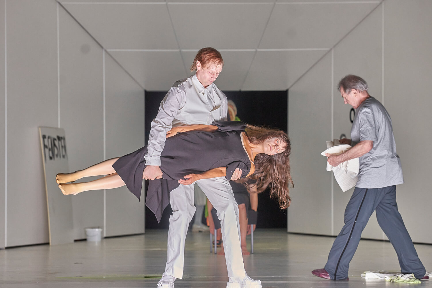 Man sieht ein Szenenfoto von „Liebe (Amour)“ von den Salzburger Festspielen. Im Vordergrund trägt ein Mann in grauer Kleidung eine Frauenpuppe, die ein schwarzes Kleid anhat. Rechts geht ein älterer Mann über die Bühne.