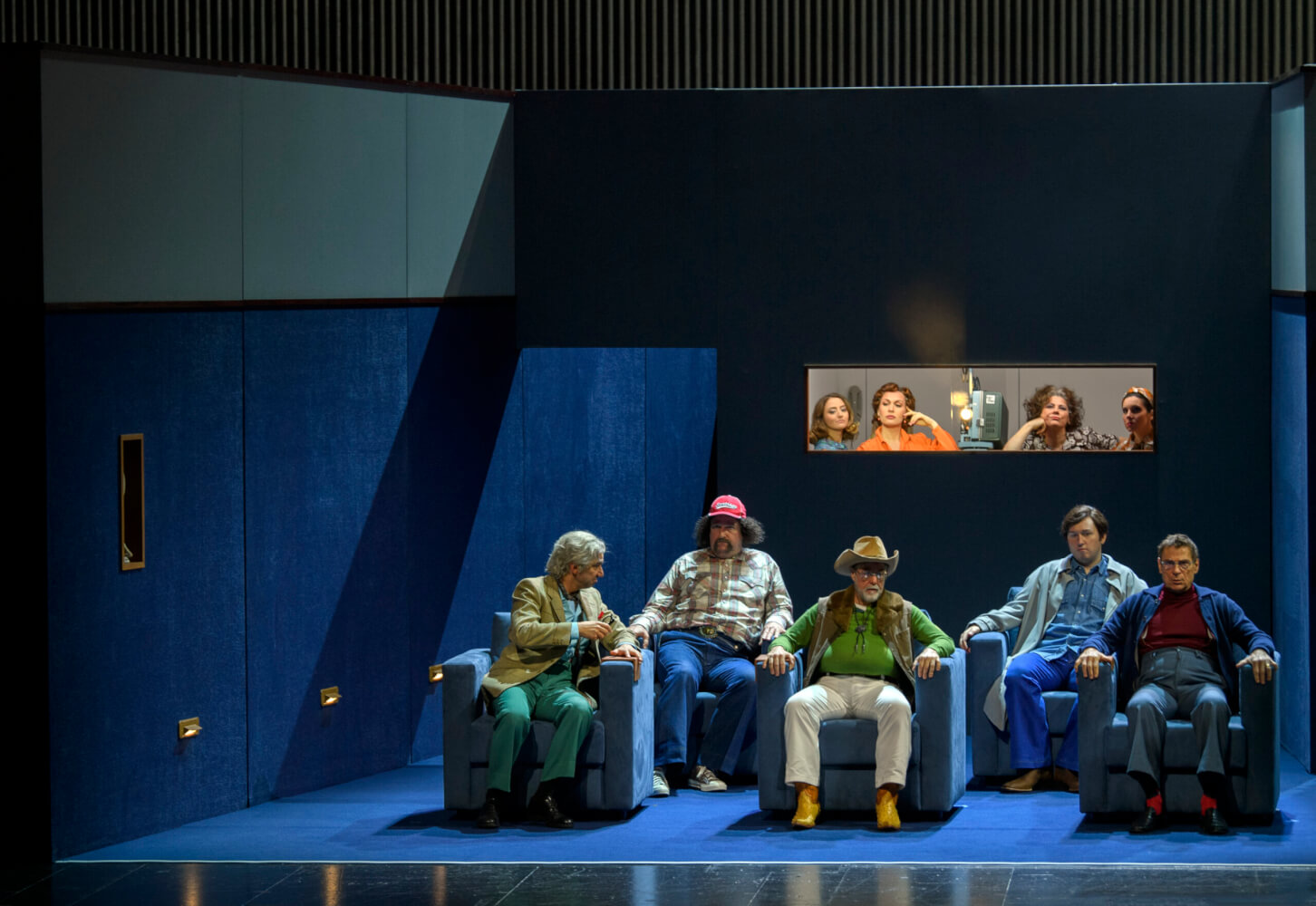 Man sieht ein Aufführungsfoto aus Verdis „Falstaff“ von den Salzburger Festspielen. Auf der Bühne sitzen fünf Männer unterschiedlichen Alters in blauen Sofasesseln. Im Hintergrund werden sie durch ein schmales Fenster von vier Frauen beobachtet.