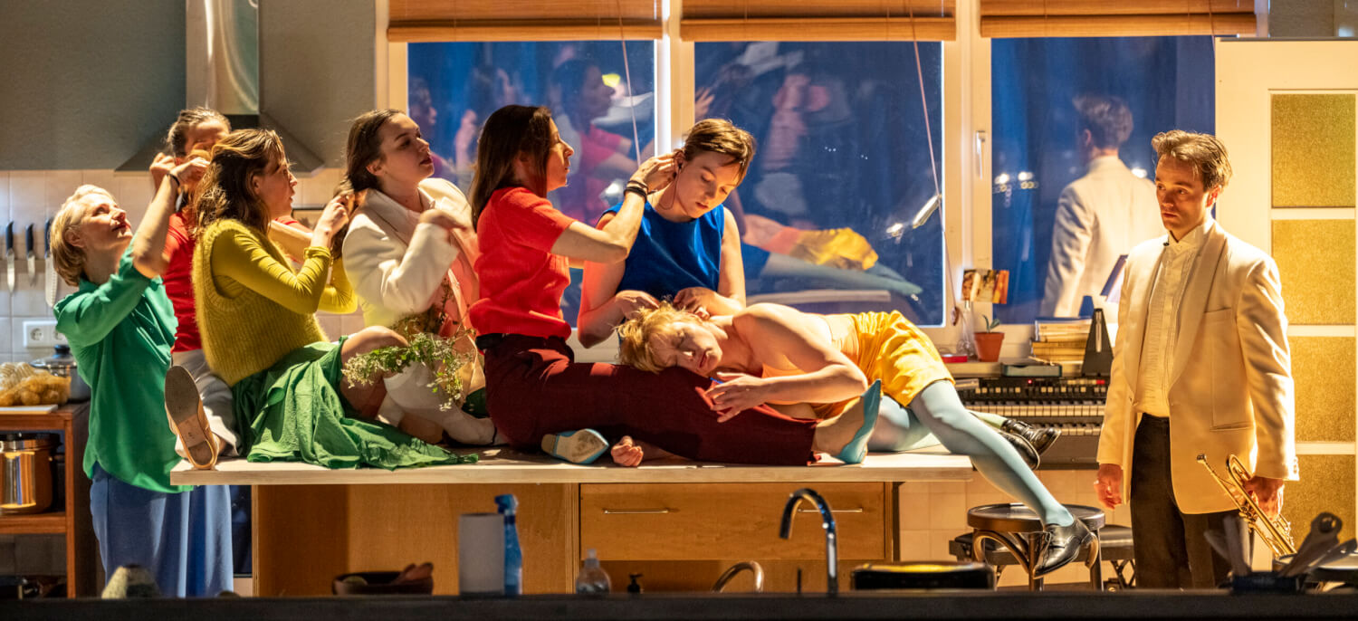 Auf der Bühne ist eine Küche eingerichtet. Das Ensemble sitzt ineinander verschränkt auf dem Küchentisch.