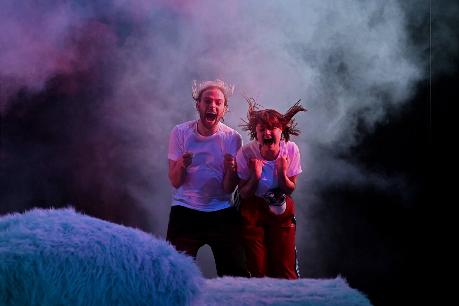 Zwei Personen stehen inmitten bunter Nebelschwaden. Von vorne kommt Wind. Sie schreien ihm entgegen.