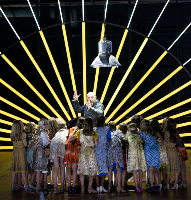 Zu sehen ist ein Szenenfoto aus Verdis „Macbeth“ von den Salzburger Festspielen. Eine Kinderschar steht im Halbkreis mit dem Rücken zum Publikum einem Mann in der Mitte zugewandt.