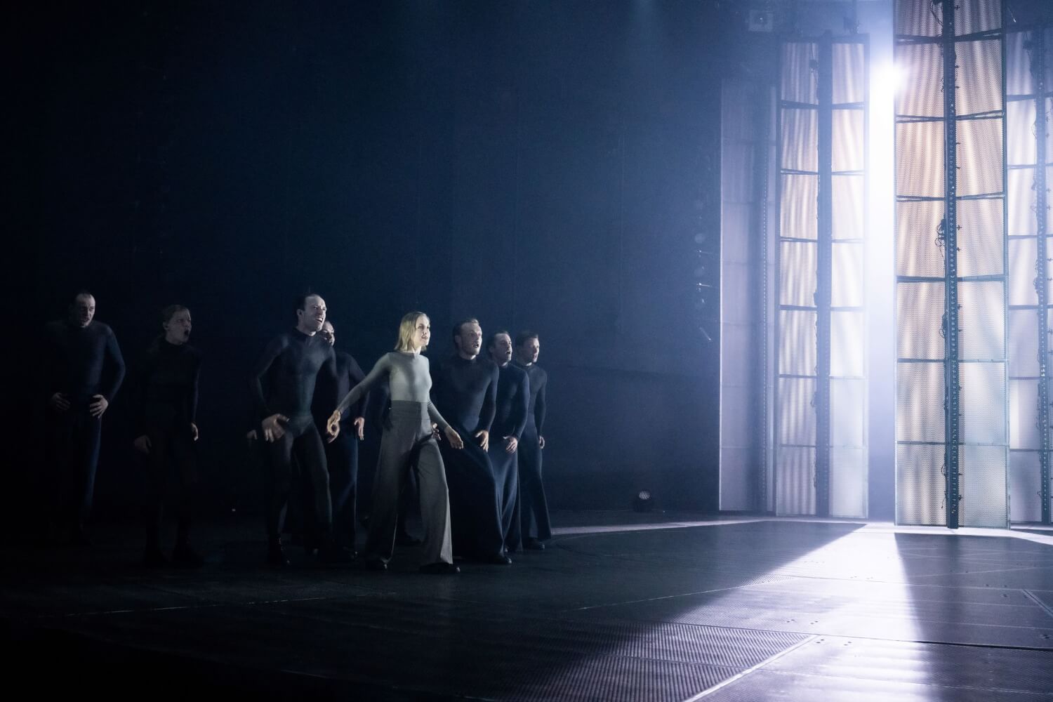 Zu sehen ist ein Szenenfoto aus Lessings „Nathan der Weise“ von den Salzburger Festspielen. Rechts öffnet sich eine hell erleuchtete Fensterfront, links wenden sich eine Frau und sieben Männer aus dem Dunkel dem Licht zu.