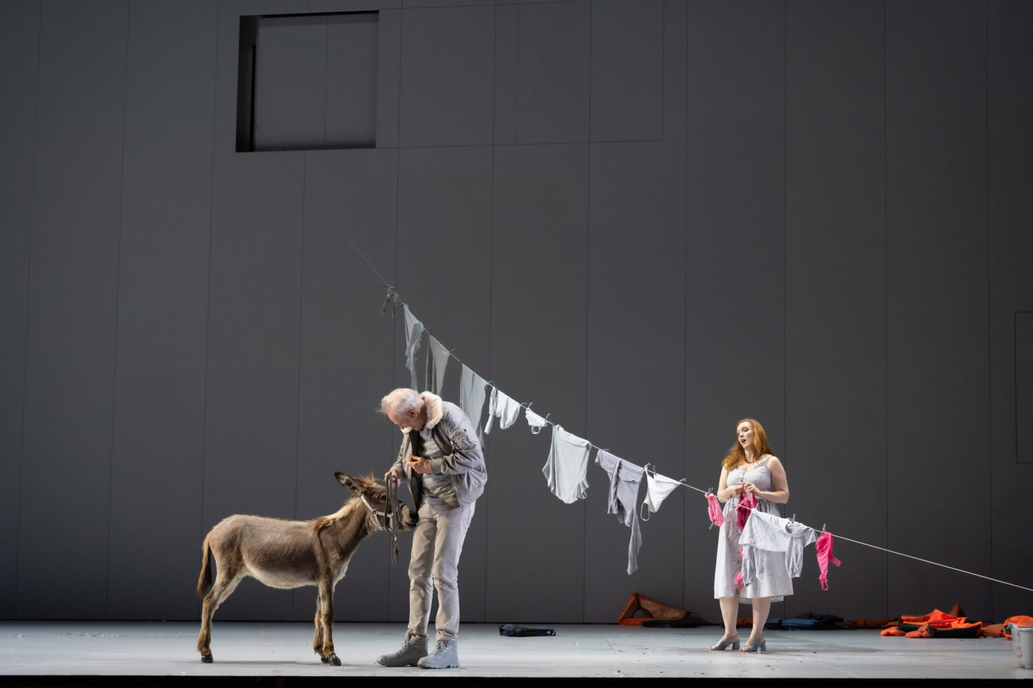 Zu sehen ist ein Aufführungsfoto von Bohuslav Martinůs „The Greek Passion“ von den Salzburger Festspiele. Vor einer grauen Bühnenwand steht ein älterer Mann, der einen Esel an der Leine führt. Im Hintergrund hängt eine jüngere Frau Kleidungsstücke auf eine Wäscheleine.