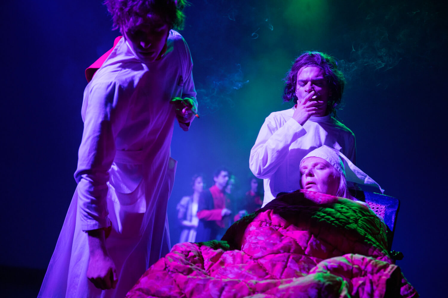 Eine schlafende, ältere Dame sitzt rechts im Vordergrund. Hinter ihr stehen zwei in weiße Kittel gekleidete Personen, die rauchen.